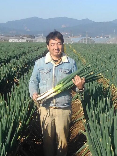 alt="みやび農園代表の林竜也。収穫したばかりの立派なねぎを手に笑顔で立つ姿"